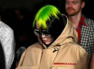 Billie Eilish wearing a beige Prada hooded jacket and oversized sunglasses, with neon green and black hair, photographed at a fashion event with dim background lighting