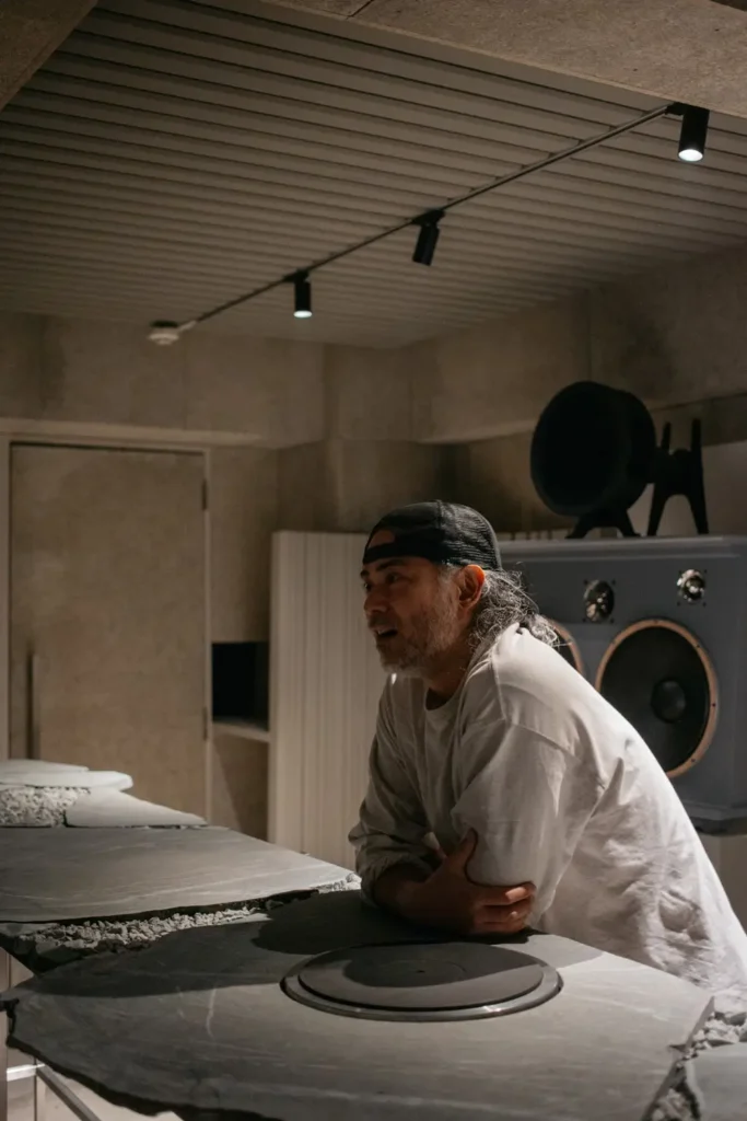 Keiichiro Oshiro inside PASS listening bar leaning over the stone turntable table with speakers behind him