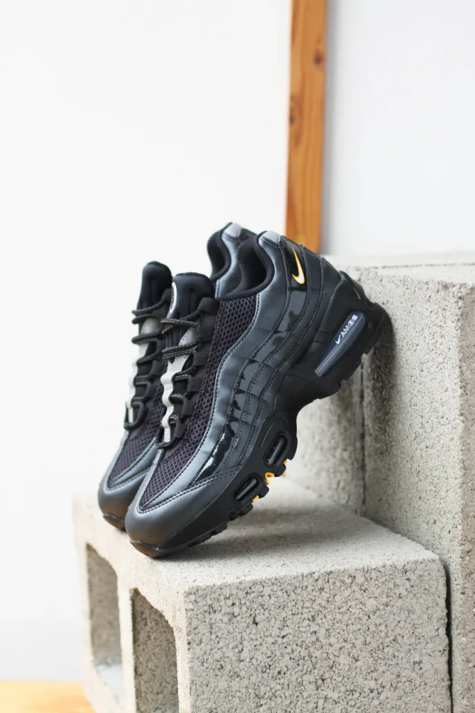 Nike Air Max 95 “Black / White – University Gold” sneakers displayed on stacked concrete blocks, featuring black mesh upper, glossy layered panels, visible Air Max cushioning, and gold Swoosh accents