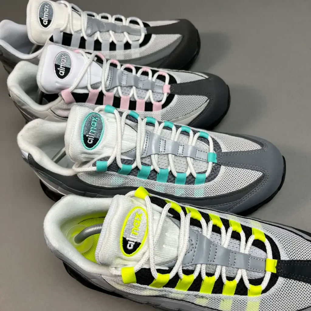 Top-down view of multiple pairs of Nike Air Max 95 sneakers in grey and white mesh with varied accent colors—including neon yellow, teal, and pink—highlighting their signature lace loops, padded tongues, and layered panel construction against a neutral surface