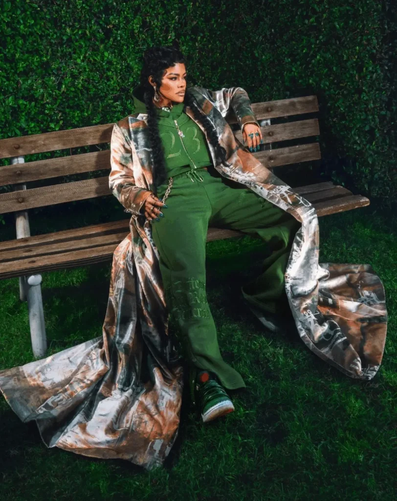 Teyana Taylor sitting on a wooden park bench wearing a green tracksuit and long patterned coat while showcasing the Teyana Taylor x Air Jordan 3 SP “Concrete Rose”