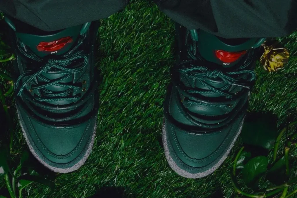 Overhead view of the Teyana Taylor x Air Jordan 3 SP “Concrete Rose” showing forest green leather construction, cement-textured mudguard, and gold rose pendant detail against a grass background