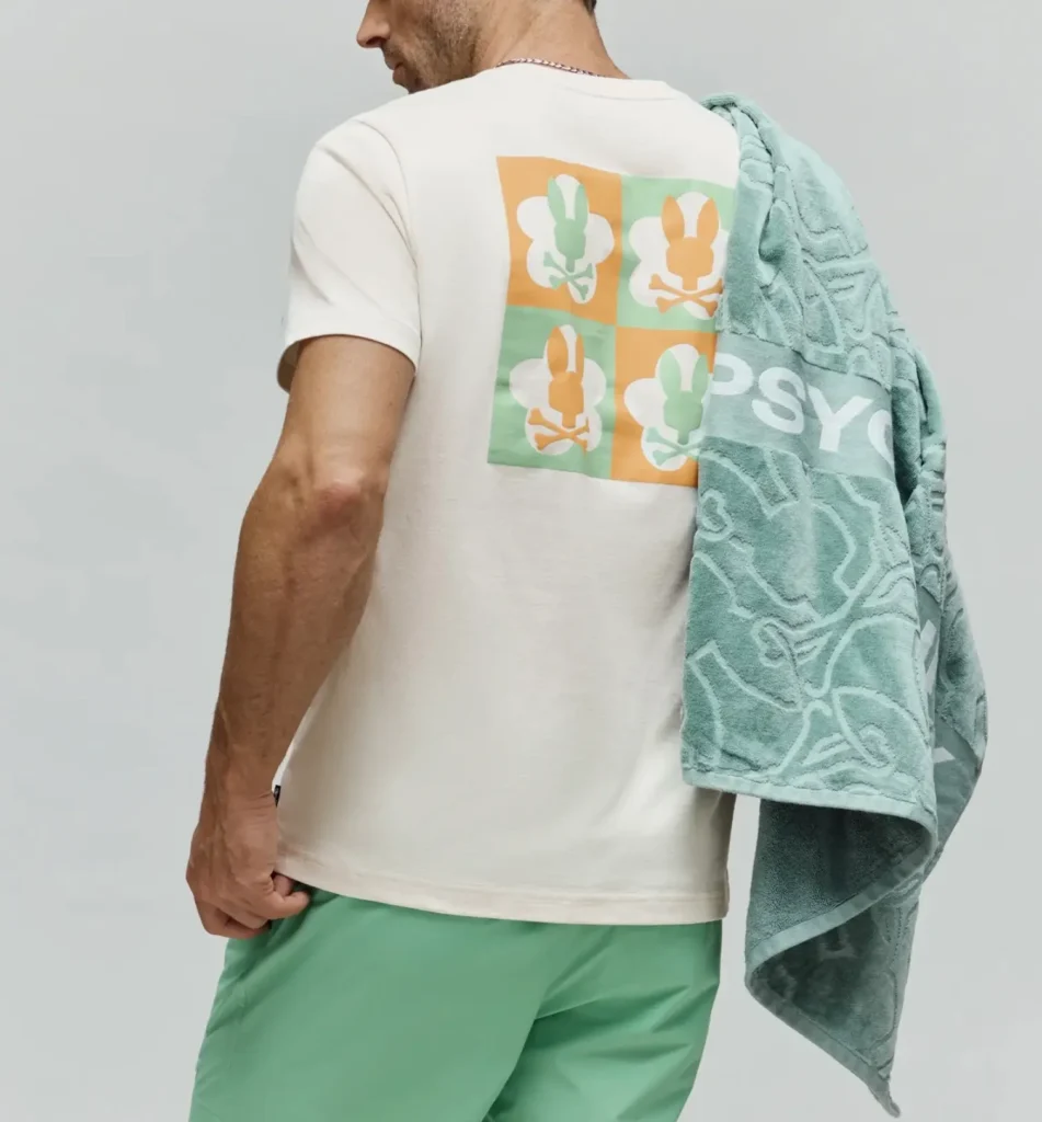 Man wearing the Psycho Bunny Men’s Aiden Back Graphic Tee in off-white featuring a large four-panel bunny skull logo graphic on the back in green and orange