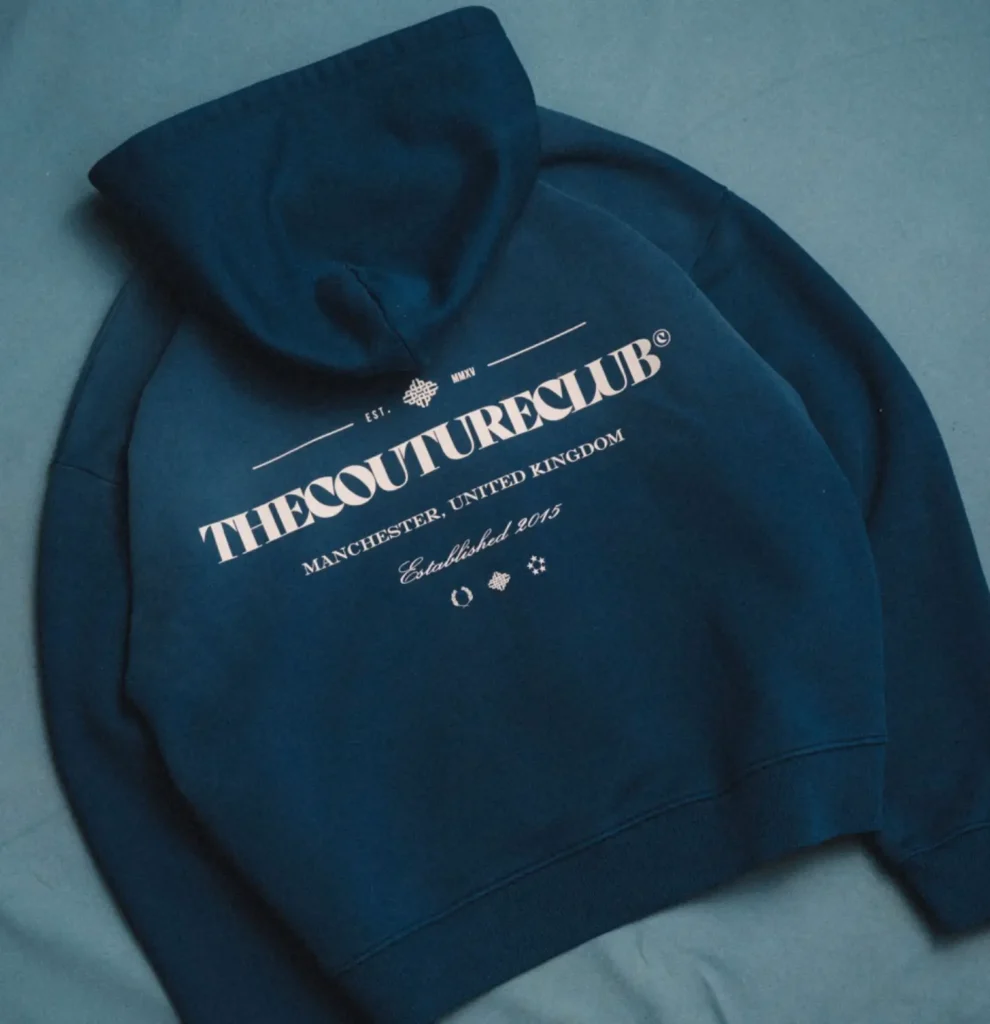 Flat lay of a navy blue The Couture Club hoodie featuring bold white logo typography, “Manchester, United Kingdom” text, and “Established 2015” detailing, highlighting soft fabric and minimalist streetwear branding