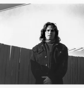 Black-and-white portrait of Jim Morrison standing outdoors in Laurel Canyon, wearing a dark jacket and scarf, framed against a wooden fence with a calm, introspective expression