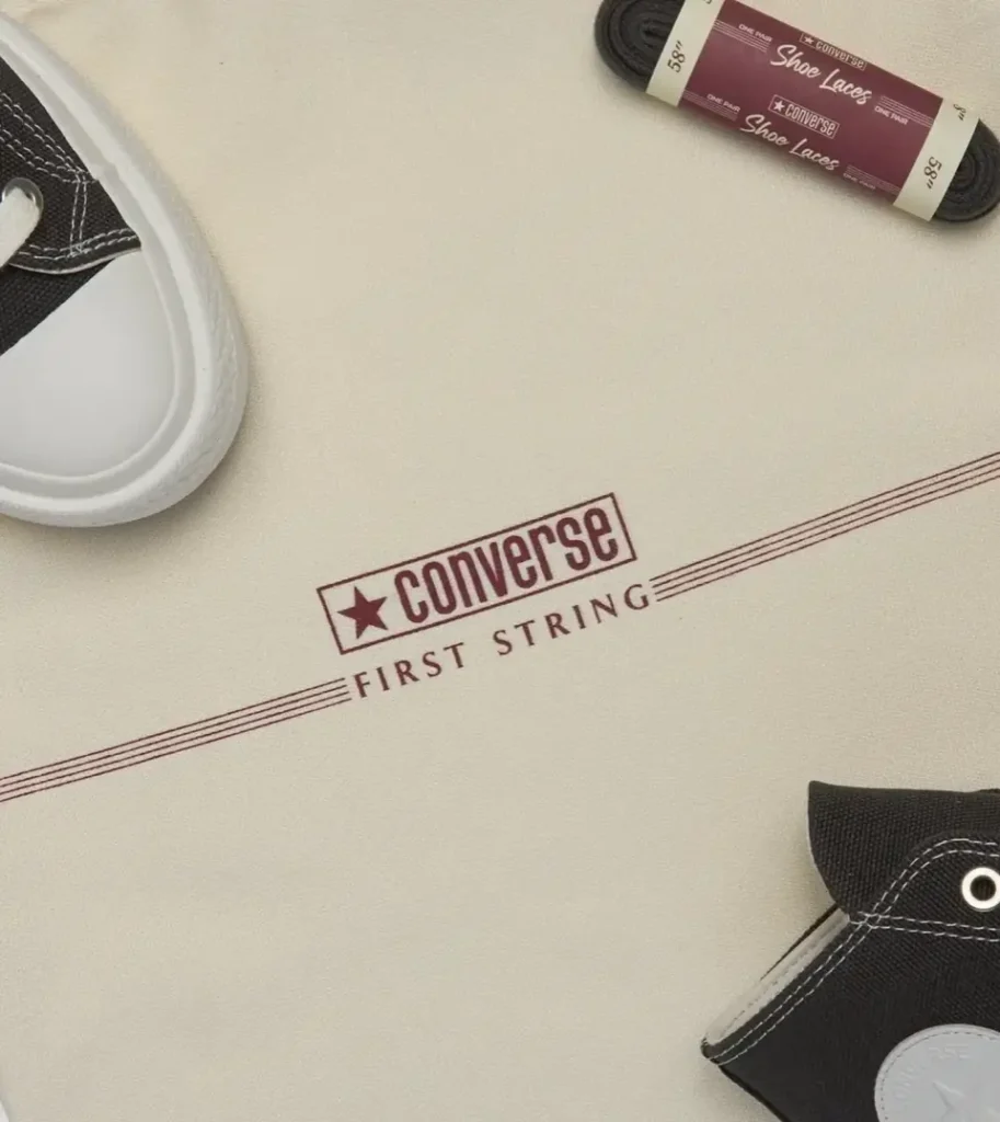 Flat-lay of Converse First String packaging on natural canvas background, featuring burgundy “Converse First String” logo, partial black Chuck Taylor high-top sneakers, and a wrapped pair of 58-inch Converse shoe laces
