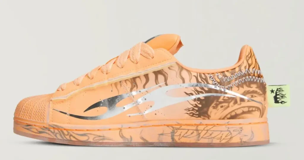 Side profile of the Hellstar x adidas Superstar ‘Hazy Orange’ featuring a soft peach-orange upper with metallic silver flame graphic overlay, tonal laces, classic shell toe, and illustrated artwork along the heel and midsole, finished with exposed stitching and a neon green branded heel tab