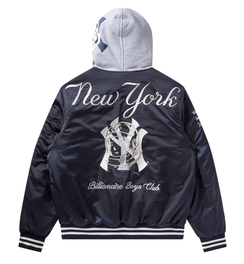 Back view of a navy satin varsity jacket from the Billionaire Boys Club x New York Yankees collaboration, featuring large script “New York” lettering, a central BBC astronaut graphic, and “Billionaire Boys Club” branding, layered with a grey hood and finished with striped ribbed hem and cuffs