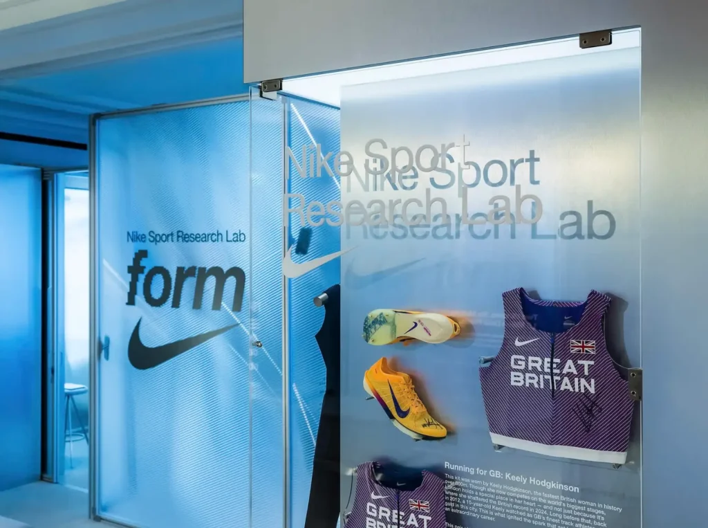 Glass display installation inside Nike Sports Research Lab pop-up featuring “Nike Sport Research Lab” branding, with mounted track spikes and a Great Britain racing singlet associated with athlete Keely Hodgkinson, set against frosted panels and layered lab-style interiors