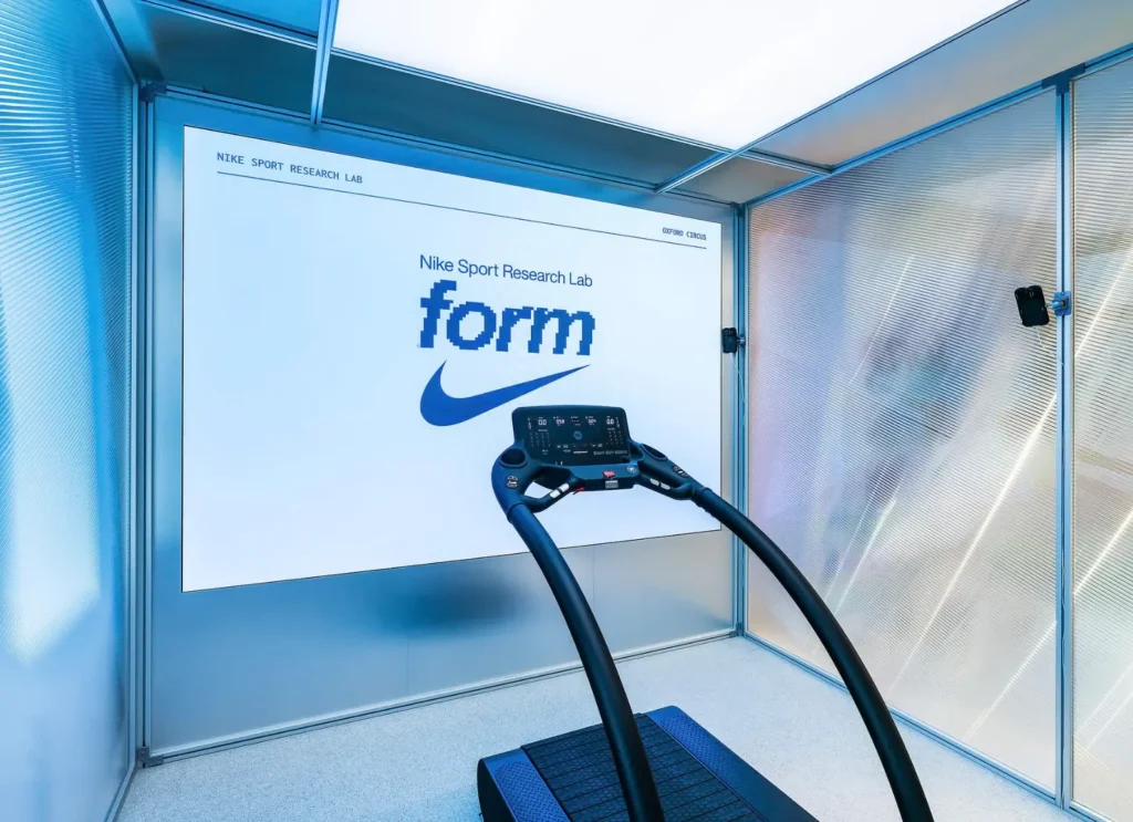 Curved treadmill positioned within Nike Sports Research Lab pop-up at Oxford Circus, facing a large display wall featuring the NSRL “form” branding, surrounded by frosted metallic panels and integrated motion-capture sensors in a controlled, laboratory-like retail space