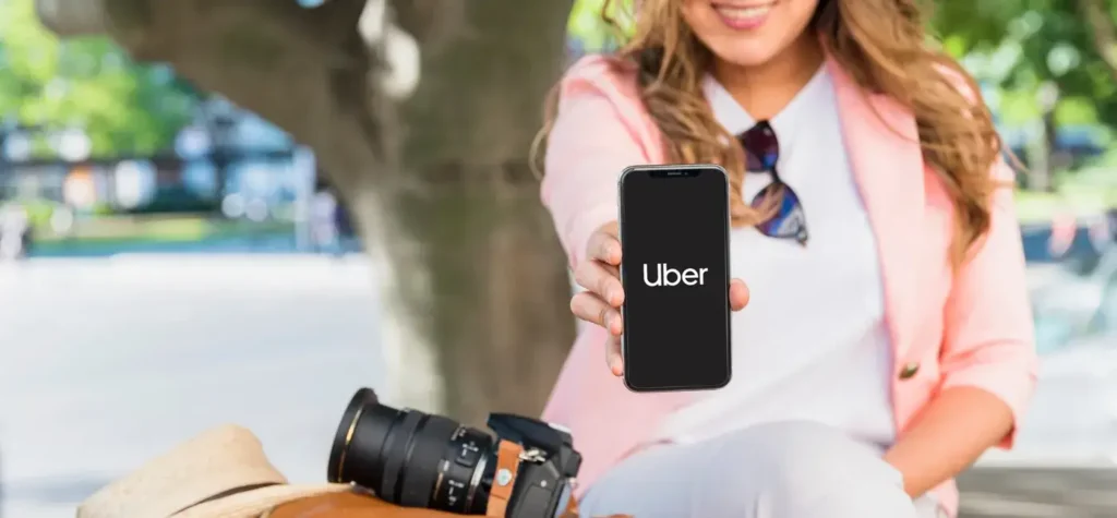 Woman holding a smartphone displaying the Uber app logo while sitting outdoors, with a camera resting beside her, illustrating the use of the rideshare platform on a mobile device