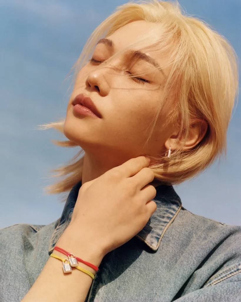Close-up portrait of a blonde model wearing Louis Vuitton Silver Lockit bracelets on red and yellow cords with small padlock charms, styled with a denim jacket against a soft blue sky background