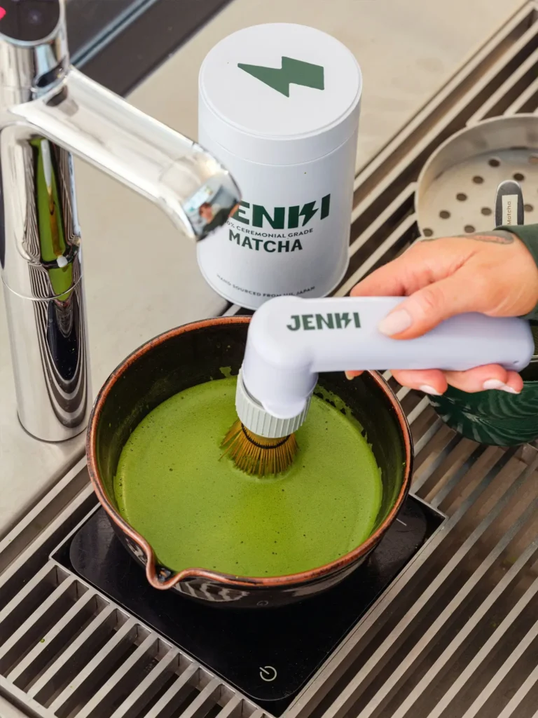 Close-up of matcha being whisked in a ceramic bowl using a handheld electric whisk, next to a container labeled Jenki ceremonial grade matcha, demonstrating modern preparation of powdered green tea in a café setting