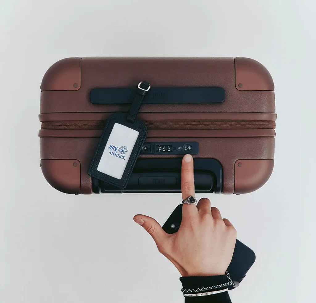 Hand pressing the built-in tracking button on a brown July CaseSafe smart suitcase, featuring an integrated lock system and luggage tag, demonstrating the suitcase’s tracking technology for locating lost baggage