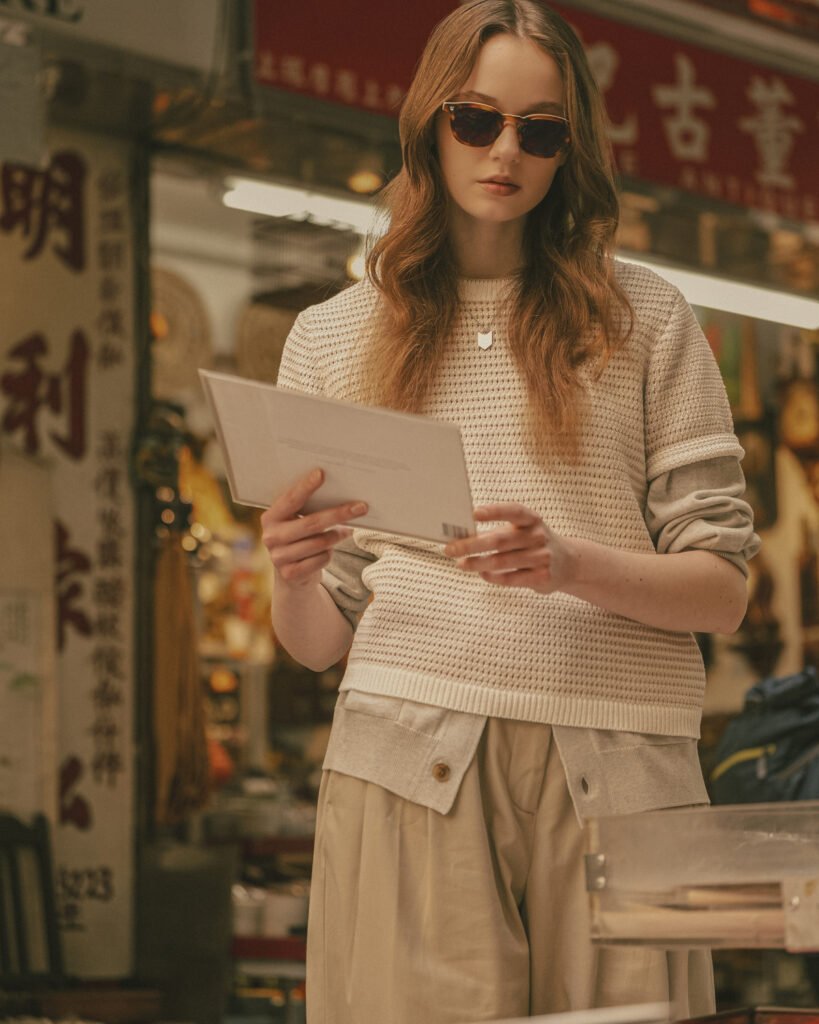 Model wearing tortoiseshell sunglasses and a cream textured knit top, holding a postcard in a Hong Kong street market setting with Chinese signage in the background, styled for A. SOCIETY × Maison Kitsuné eyewear campaign
