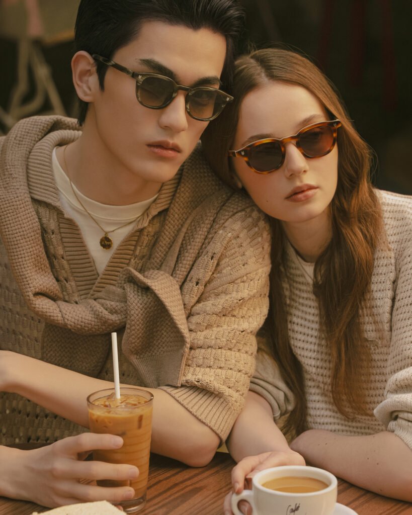 Two models wearing tortoiseshell and dark acetate sunglasses sit closely at a café table with drinks, styled in neutral knitwear, showcasing the A. SOCIETY × Maison Kitsuné eyewear capsule in a warm, lifestyle setting