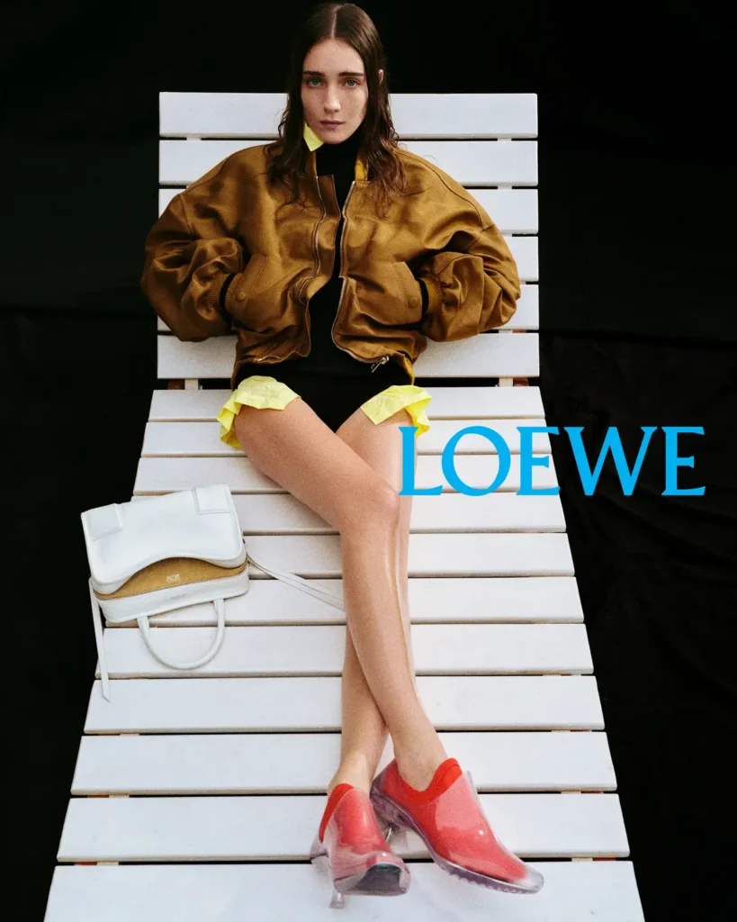 Model seated on a white lounge chair wearing a brown bomber jacket and red shoes, with a white Loewe Amazona 180 bag placed beside her in the Loewe Spring/Summer 2026 campaign