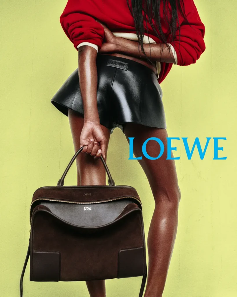 Model wearing a red sweater and black leather skirt holding a brown Loewe Amazona 180 bag with top handle against a lime-green background in the Loewe Spring/Summer 2026 campaign