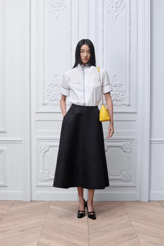 Full look featuring a model wearing the Small Dior Bow Bag in buttercup yellow lambskin with a gold-tone chain strap, styled with a white short-sleeve blouse, black midi skirt, and black heels against an ornate white interior backdrop