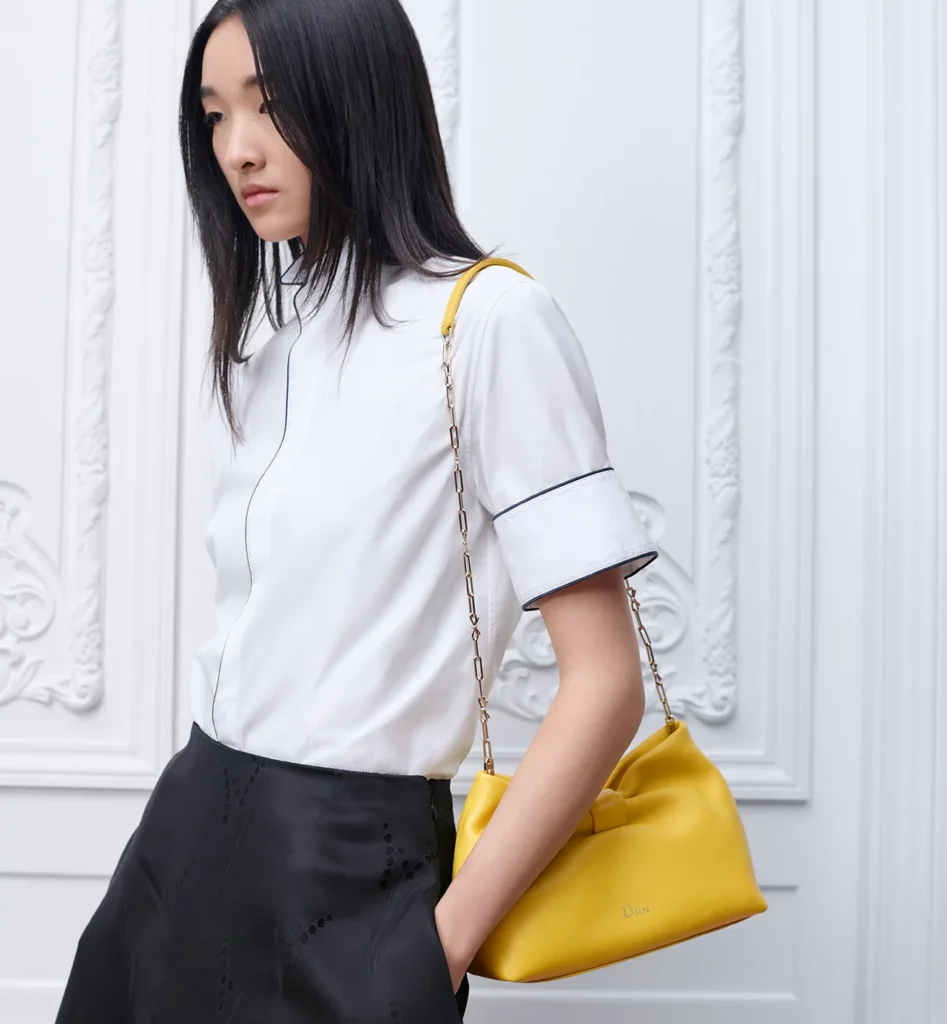 Model wearing the Small Dior Bow Bag in buttercup yellow lambskin with a gold-tone chain shoulder strap, styled with a white blouse and black skirt in a minimal interior setting