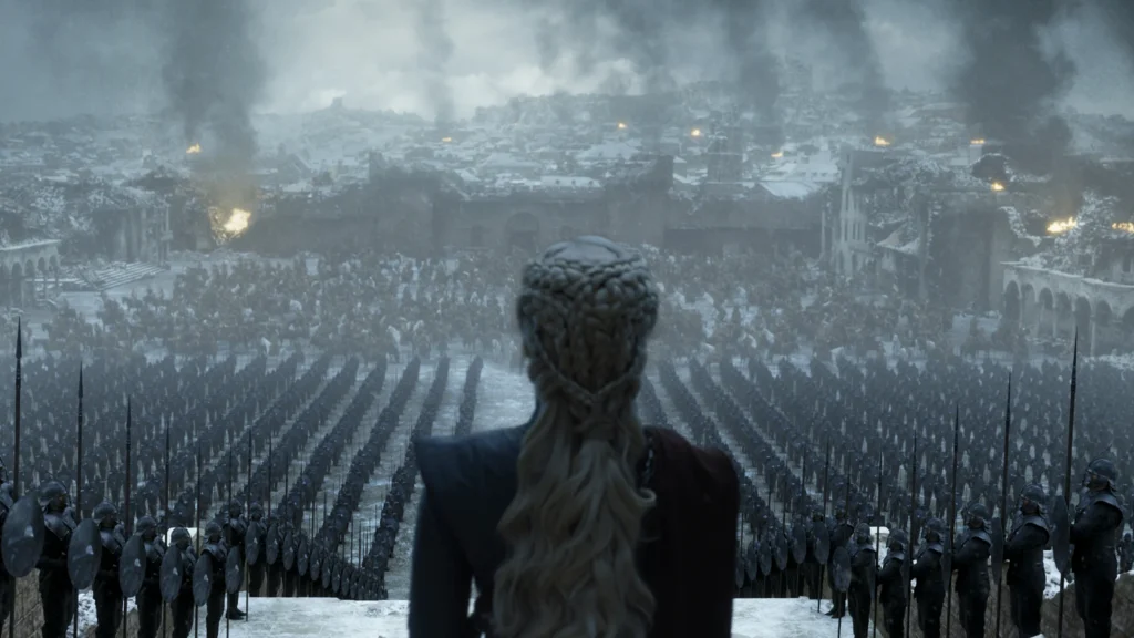 Daenerys Targaryen stands before a massive formation of soldiers in a snowy battlefield outside a ruined city in a dramatic scene from Game of Thrones, as smoke rises over the crowd of assembled warriors