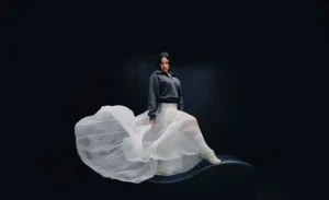 Rosalía posing against a dark studio background wearing a flowing white skirt and gray top while showcasing the New Balance 204L shoes during the brand’s campaign shoot