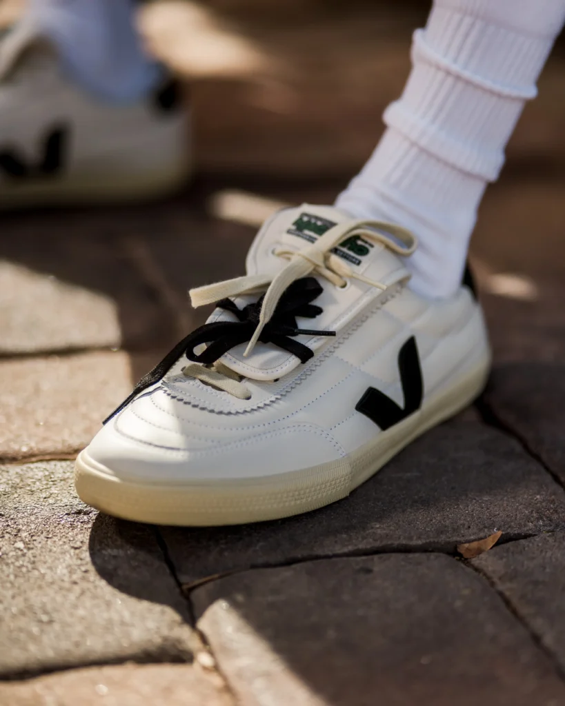 Magliano x VEJA Panenka shoe in white leather with black V logo, featuring a fold-over football-style tongue and mixed-color laces worn with white ribbed socks on a stone pavement