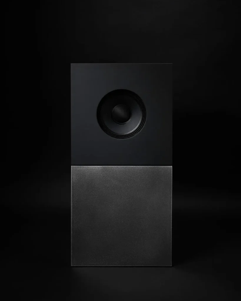 Cubist-inspired minimalist speaker from the Nocs Braque stereo system by Daniel Alm, featuring a matte black cube with a centered circular driver stacked atop a brushed-metal rectangular base against a dark background
