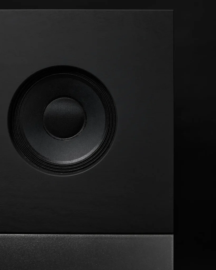Close-up of a Nocs Braque stereo speaker by Daniel Alm showing a circular driver set within a matte black cube enclosure above a brushed-metal base against a dark background
