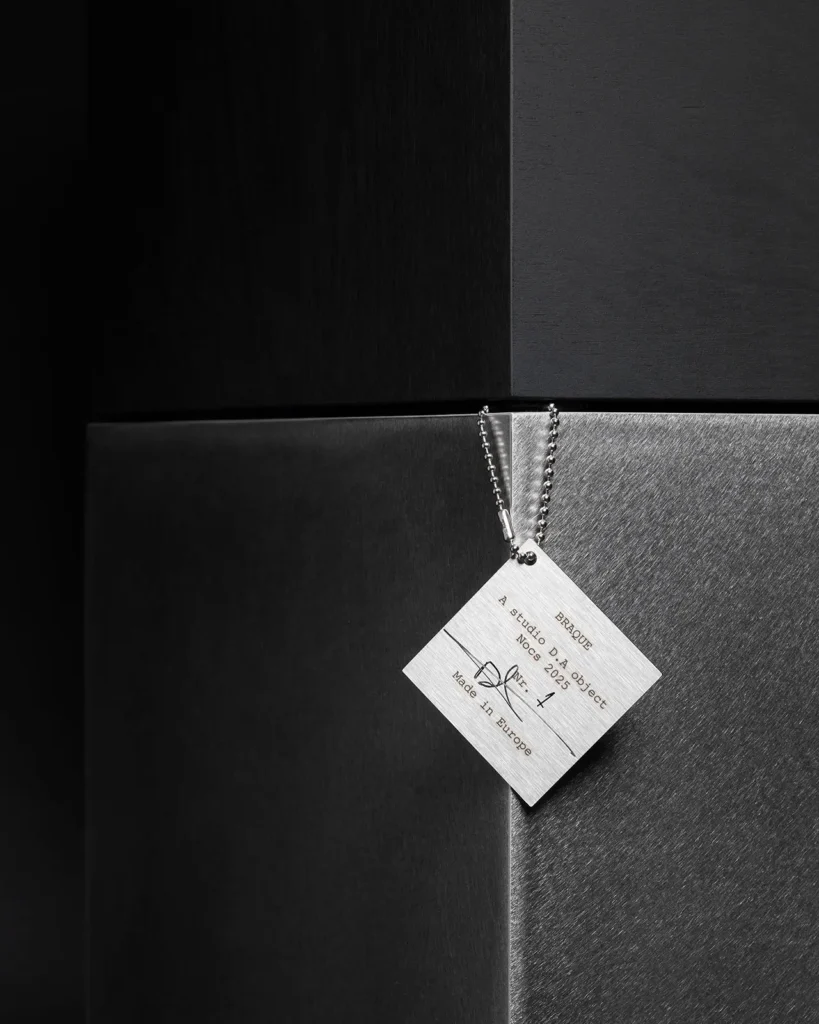 Close-up detail of the Nocs Braque stereo system by Daniel Alm showing brushed-metal cube surfaces and a hanging metal tag engraved “Braque – A Studio D.A object, Nocs 2025, Made in Europe.”