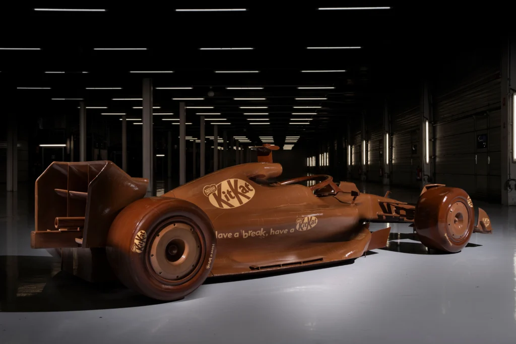 Life-sized chocolate Formula 1 race car branded with KitKat logos displayed in a dark exhibition space as part of a promotional stunt celebrating Formula One motorsport culture