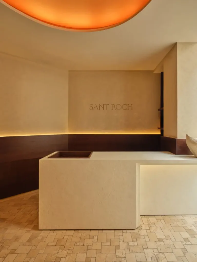 Minimalist reception area at SANT ROCH wellness space in Paris, featuring a sculptural stone desk, warm ambient lighting, and the SANT ROCH logo on a textured plaster wall beneath a glowing circular ceiling light