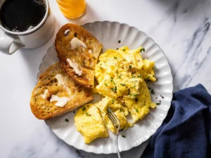 Creamy soft scrambled eggs topped with chives and black pepper served on a white plate with buttered toasted sourdough, alongside a cup of black coffee and orange juice on a marble breakfast table