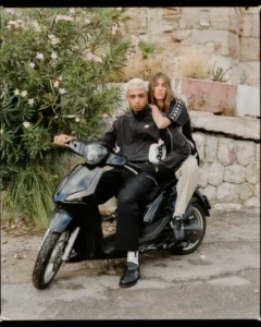Lifestyle image of two models posed on a black scooter, styled in coordinated looks from Kappa. The front rider wears a black tracksuit with subtle piping and a small Omini logo, while the second model pairs a black top with signature Banda logo taping along the sleeves and light-toned trousers. Set against a rustic stone wall and overgrown greenery, the scene carries a Mediterranean street feel—casual yet composed. The scooter adds a sense of motion and everyday utility, reinforcing the collection’s blend of sport heritage, urban mobility, and relaxed lifestyle dressing