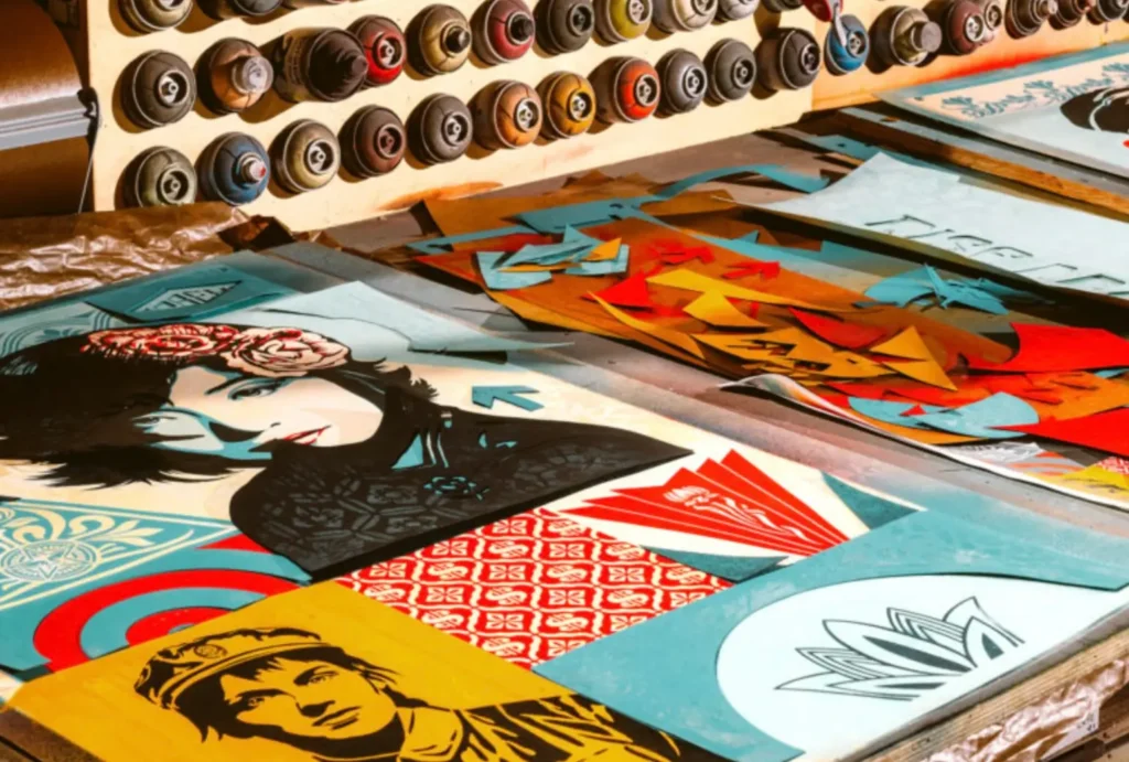 A Shepard Fairey studio worktable filled with colorful stencil cutouts, spray paint cans arranged on a wall rack, and layered graphic artwork featuring stylized portraits, geometric patterns, and OBEY-inspired iconography used in the artist’s printmaking proces
