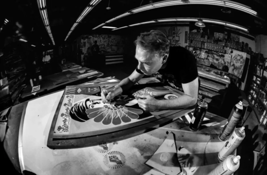 Shepard Fairey works intently in his studio, leaning over a stencil-based artwork featuring a stylized female portrait and radiating floral pattern. Spray paint cans and stencil sheets surround the workspace under curved overhead lights, capturing the hands-on process behind his iconic OBEY-style graphic compositions