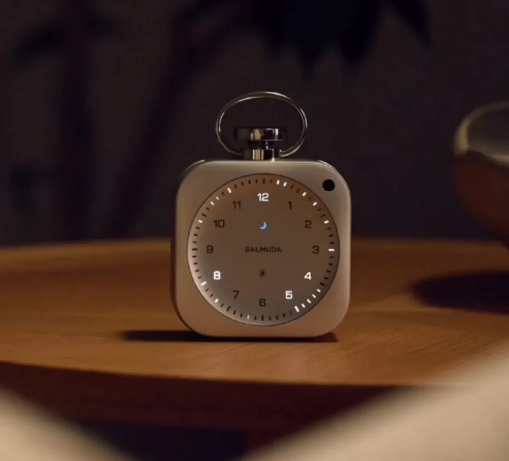 BALMUDA The Clock resting on a wooden table in a low-light, intimate setting, its softly glowing dial and subtle moon phase icon creating a warm, ambient nighttime atmosphere that emphasizes calm, sensory timekeeping