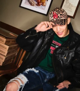 Model wearing a Dolce & Gabbana x ’47 leopard print baseball cap with red LA Dodgers logo, styled with a black leather jacket and distressed denim, seated beside stacked pizza boxes in a casual lifestyle setting