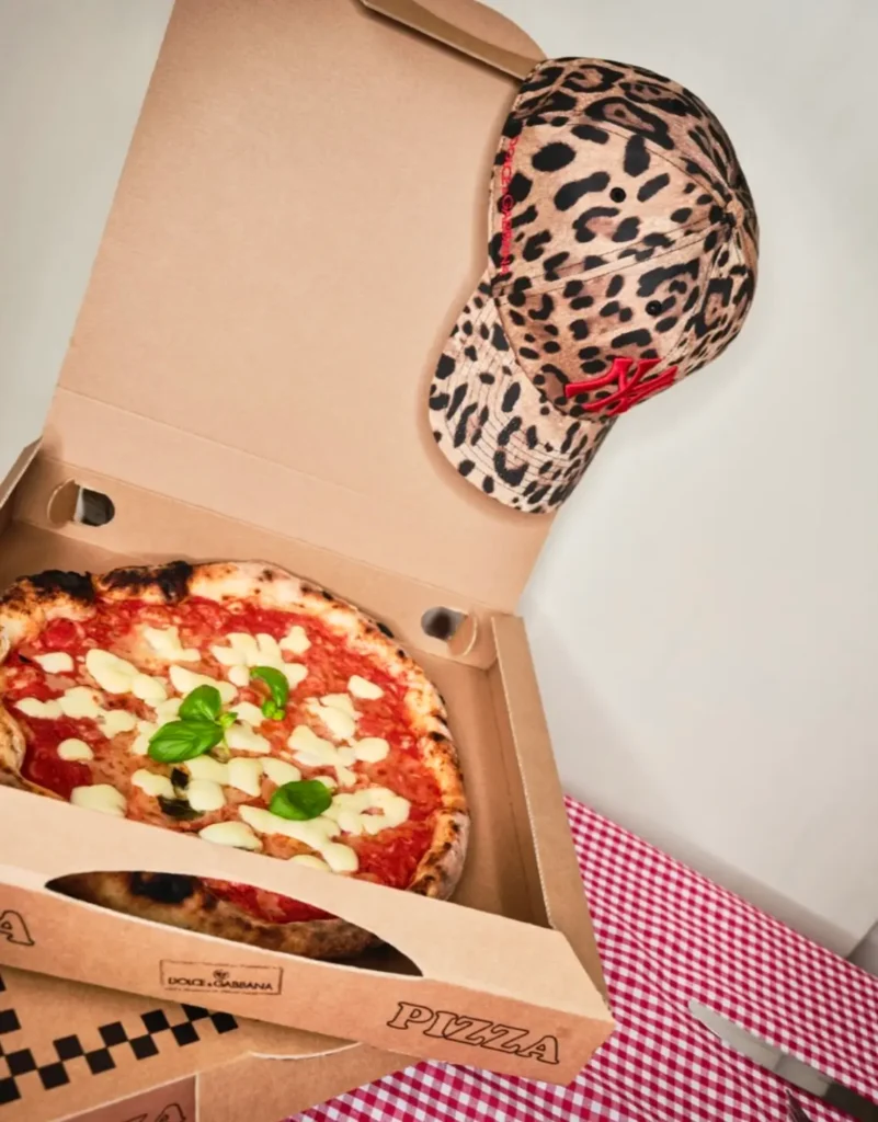 Dolce & Gabbana x ’47 leopard print baseball cap with red NY logo placed inside an open pizza box beside a Margherita pizza, styled in a playful lifestyle editorial setting with checkered tablecloth