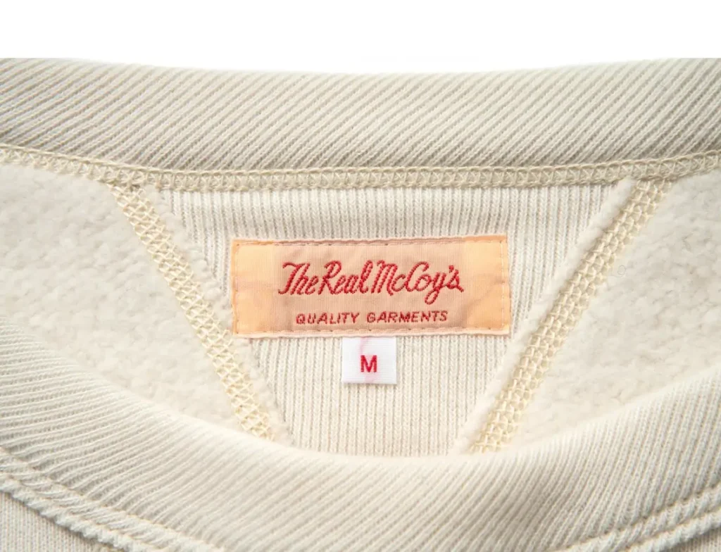 Close-up of The Real McCoy’s 10oz loopwheel crewneck sweatshirt interior showing cream cotton fleece lining, ribbed collar, triangular V-insert, and woven “The Real McCoy’s Quality Garments” label with size M tag