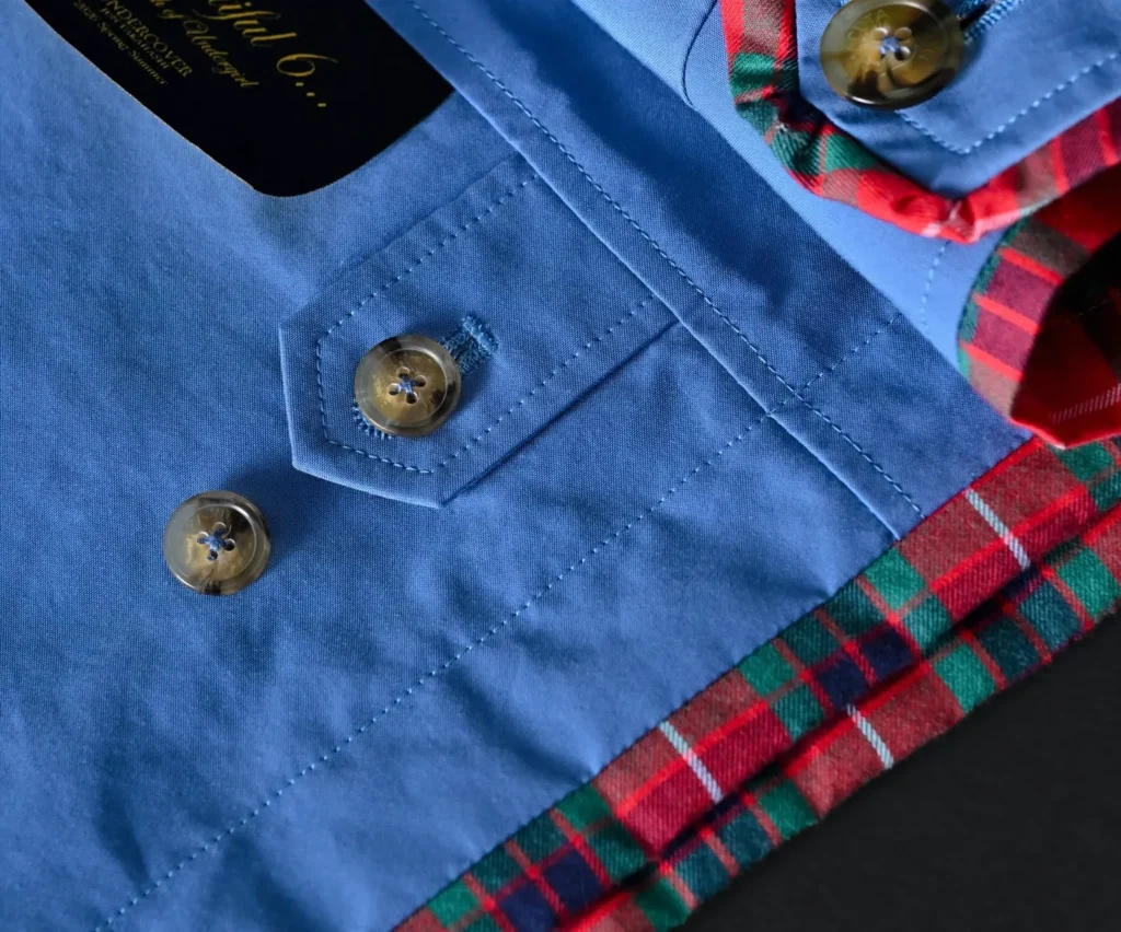 Detailed close-up of BARACUTA x UNDERCOVER swing top showing blue fabric stitching, horn-style buttons, and red tartan trim from the SS26 “but beautiful 6…” collection