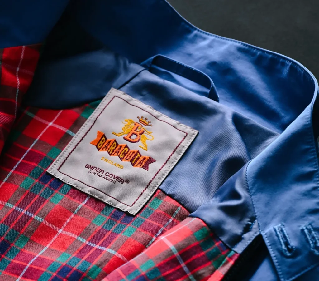 Close-up of BARACUTA x UNDERCOVER co-branded label inside blue swing top jacket, featuring red tartan lining and embroidered logo detail from the SS26 “but beautiful 6…” collection