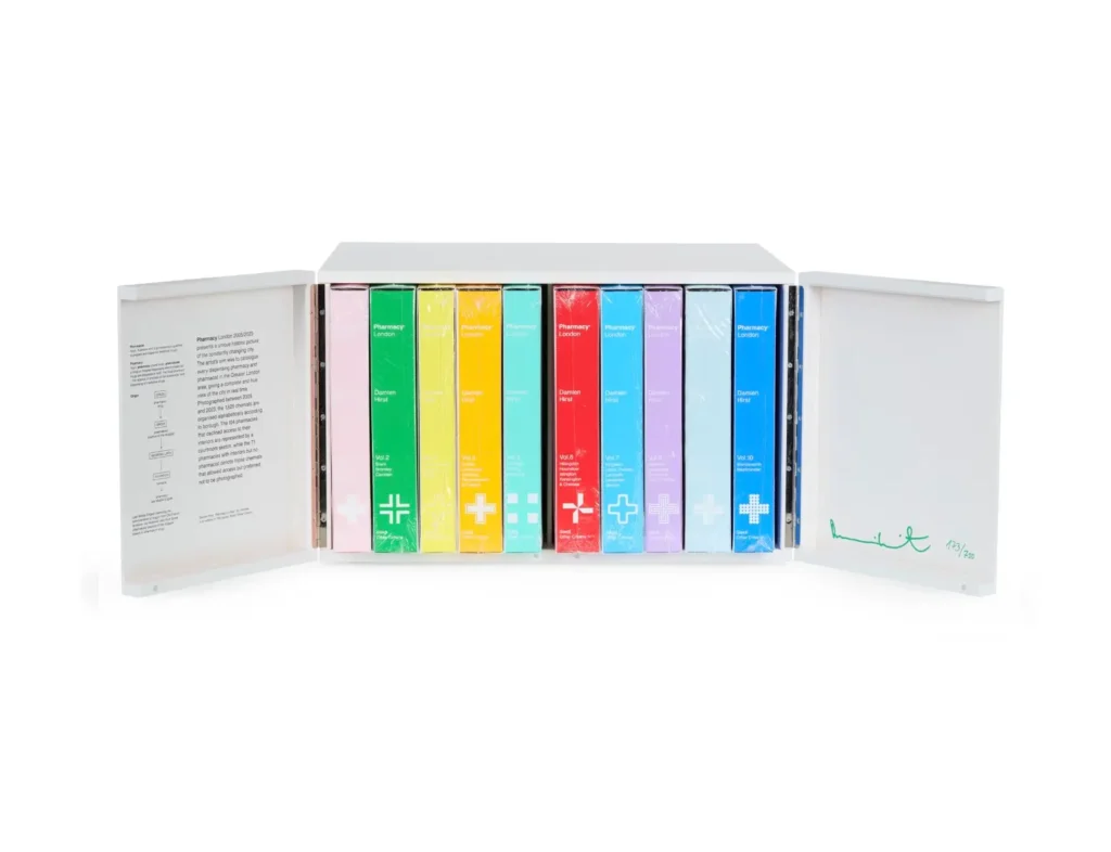 Open Damien Hirst “Pharmacy London” boxed set revealing a row of brightly colored volumes arranged like pharmaceutical packages, with white cabinet doors displaying printed text on the left and a green signature edition marking on the right