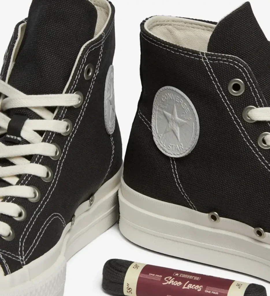 Close-up of Converse First String Chuck Taylor All Star Hi in Beluga/Black, showing textured canvas upper, contrast white stitching, metal eyelets, off-white laces, and tonal All Star ankle patch, with spare Converse 58-inch laces in packaging
