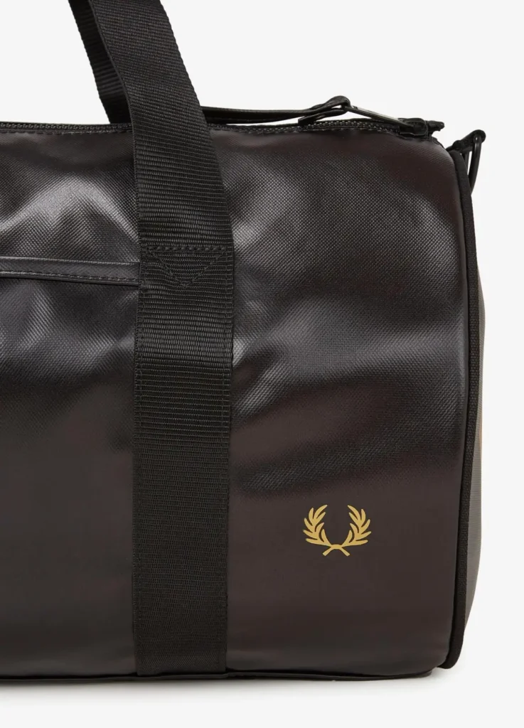Close-up of the Fred Perry Tonal Barrel Bag L7260 showing textured black coated material with subtle sheen, vertical black webbing strap stitched into the body, tonal zip closure along the top edge, and gold laurel wreath logo printed on the side panel