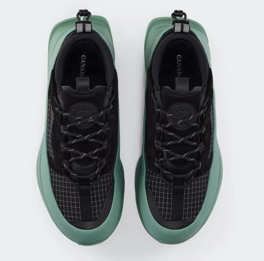 Top-down view of the Women’s Glacier Trail Sneaker in Dyneema®, showing the black ripstop grid upper, padded collar, reflective lace system, central webbing strap, and visible Canada Goose branding on the insole, framed by a sculpted sage-green rubberized rand for durability and all-terrain support