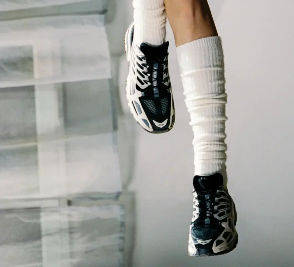 Suspended on-foot view of the Salomon ACS Pro Shell in Rainy Day/Black/Castlerock, featuring a ventilated black mesh upper with off-white cage overlays and Quicklace system, paired with ribbed white socks against a minimal interior backdrop