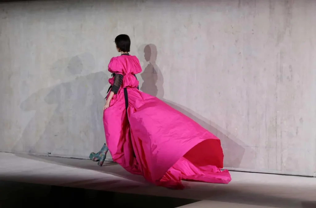 Model walking away along a minimalist runway in a voluminous bright pink gown with exaggerated structure and a sweeping train, contrasted by sheer black polka-dot sleeves and cinched detailing, creating a dramatic silhouette against a raw concrete backdrop