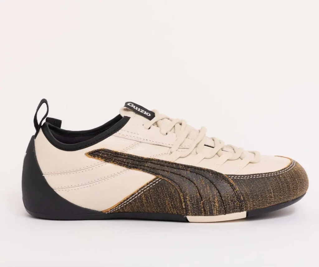 Side view of the PUMA x GUIZIO Klim Low shoe, highlighting the cream leather upper, tonal lacing, black heel counter, and textured brown formstrip overlay wrapping the toe and midfoot along the low-profile sole