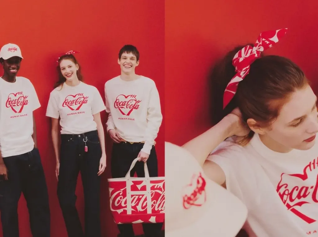 Three models posed against a vivid red backdrop wearing HUMAN MADE × Coca-Cola pieces, including white graphic T-shirts and a sweatshirt with heart-shaped logo prints, styled with a branded tote bag, cap, and a red patterned bandana for a cohesive, play streetwear look
